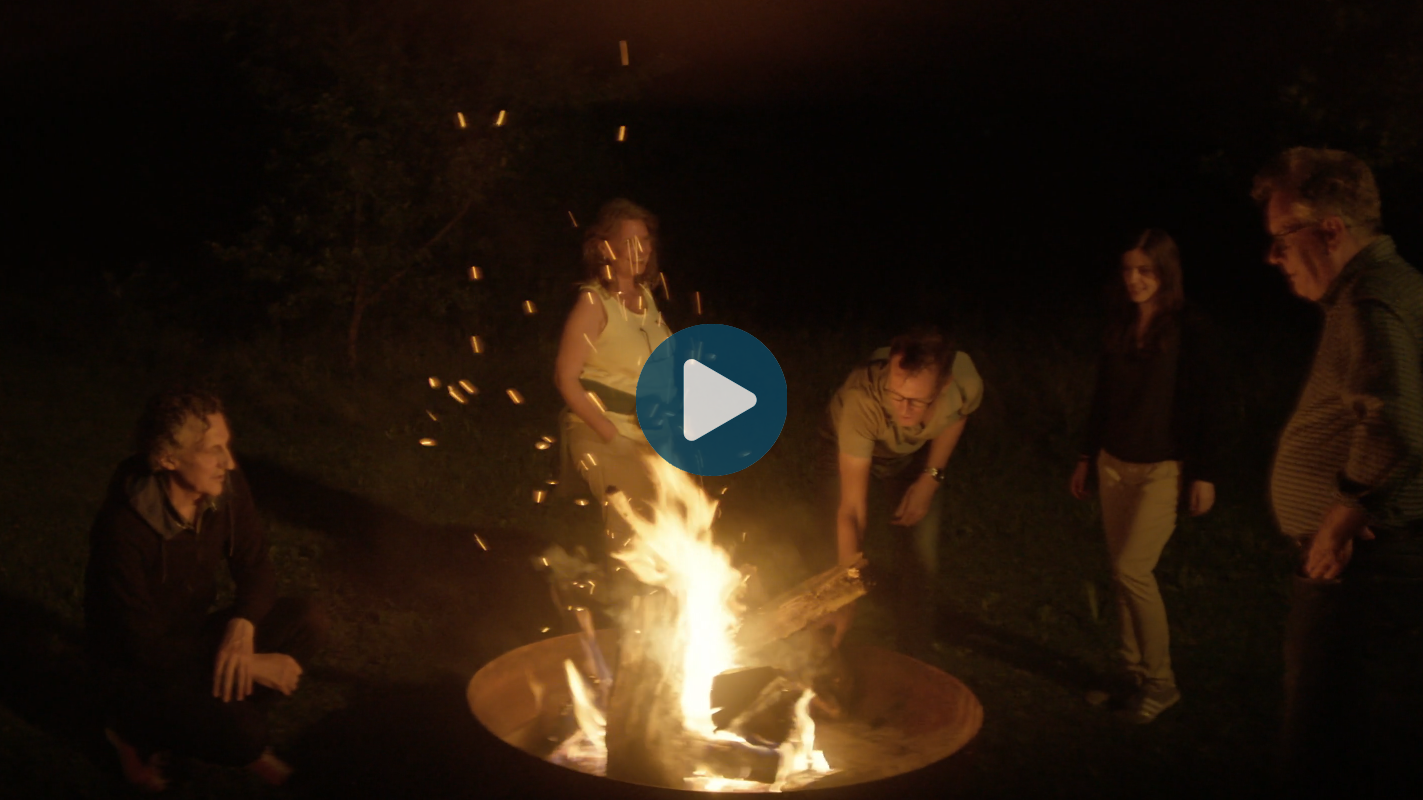 Eine Gruppe Menschen steht um ein Feuer und unterhält sich. In der Mitte des Bildes ist ein Video-Play-Button abgebildet.